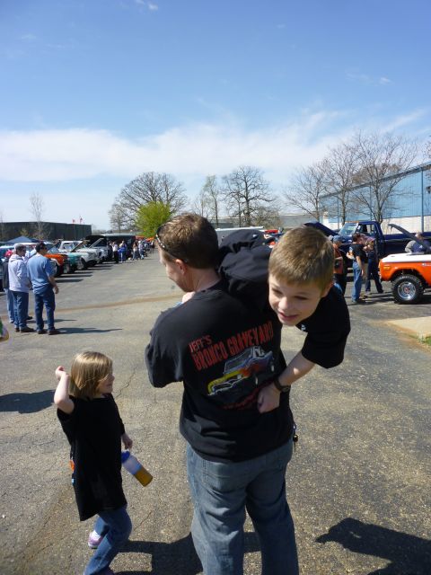 1995 Ford Bronco - A father carries his son while a young girl stands nearby at an outdoor car show with many vehicles on display.