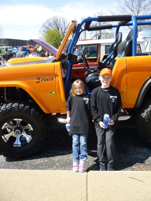 1995 Ford Bronco - A bright orange Ford Bronco with a blue roll cage is parked at an outdoor event, with two children posing in front of it.