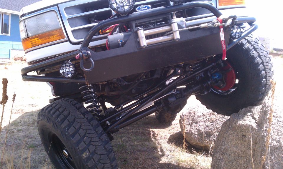 Lifted white Ford 4x4 flexing front axle on a boulder with Warn winch bumper and PIAA lights