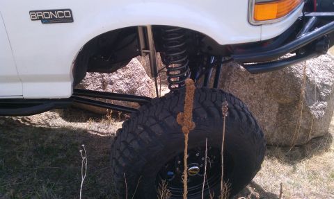 1978 Ford Bronco - A white Ford Bronco is shown with its off-road suspension and large tires, parked near rocks. The 'Bronco' badge is visible on the fender.