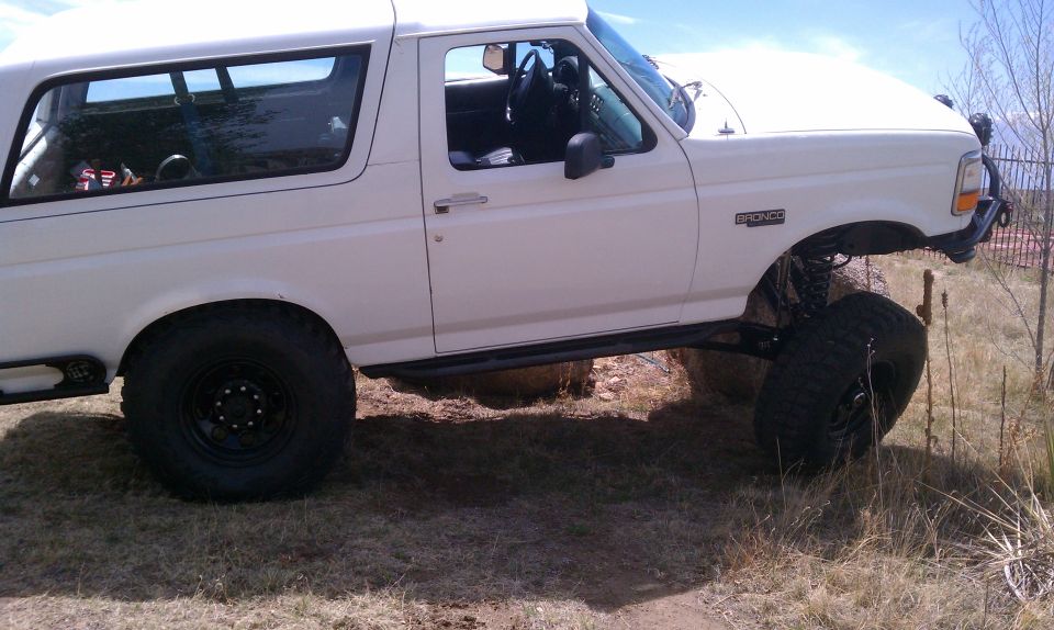 White 1990s Ford Bronco on a lift kit flexing over a dirt bank, black steel wheels and mud tires