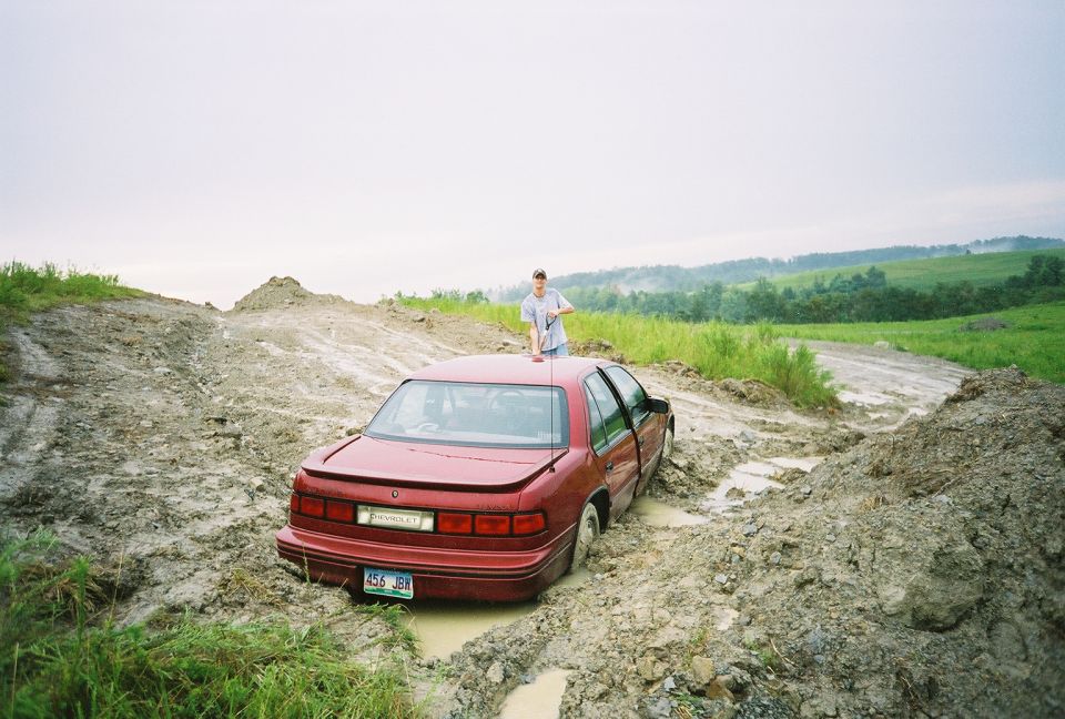 Red Chevrolet sedan stuck in deep mud on a rutted trail, man standing beside it, green hills behind