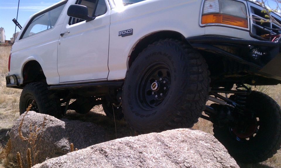 White 1990s Ford Bronco lifted on mud tires crawling over granite boulders, low front angle
