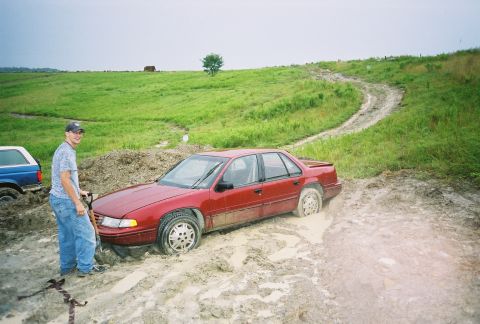 1990 Ford Bronco - Red sedan and blue SUV stuck in mud on a dirt road with grassy hills in the background.