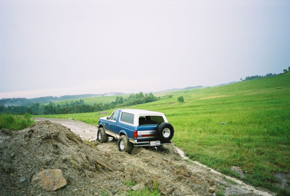 Lifted blue and white Ford Bronco with big mud tires and rear spare on a muddy trail through green fields