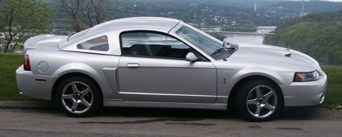 2007 Ford Mustang - A silver Ford Mustang coupe is seen from the side, parked on a road with a view of a bridge and town.