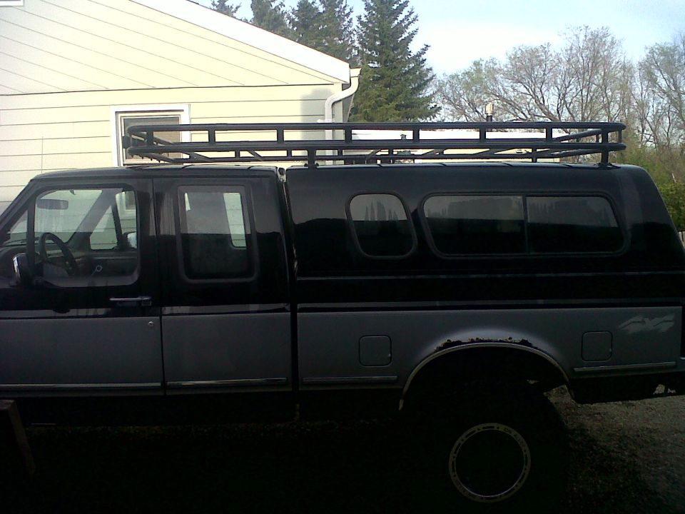 Two-tone gray and black Ford F-150 pickup truck with a black camper shell and roof rack, seen from the side