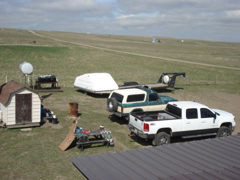 2011 Yugo Vega - A rural property features two pickup trucks, a trailer with a camper shell, and a small shed under a cloudy sky.