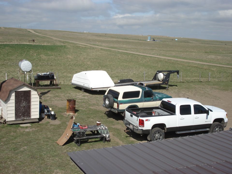 White Z71 crew-cab pickup and green and tan SUV parked in a prairie farmyard with trailer and shed
