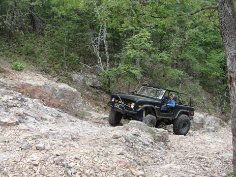 1967 Ford Mustang - Black Ford Bronco navigates a challenging rocky trail in a wooded area.