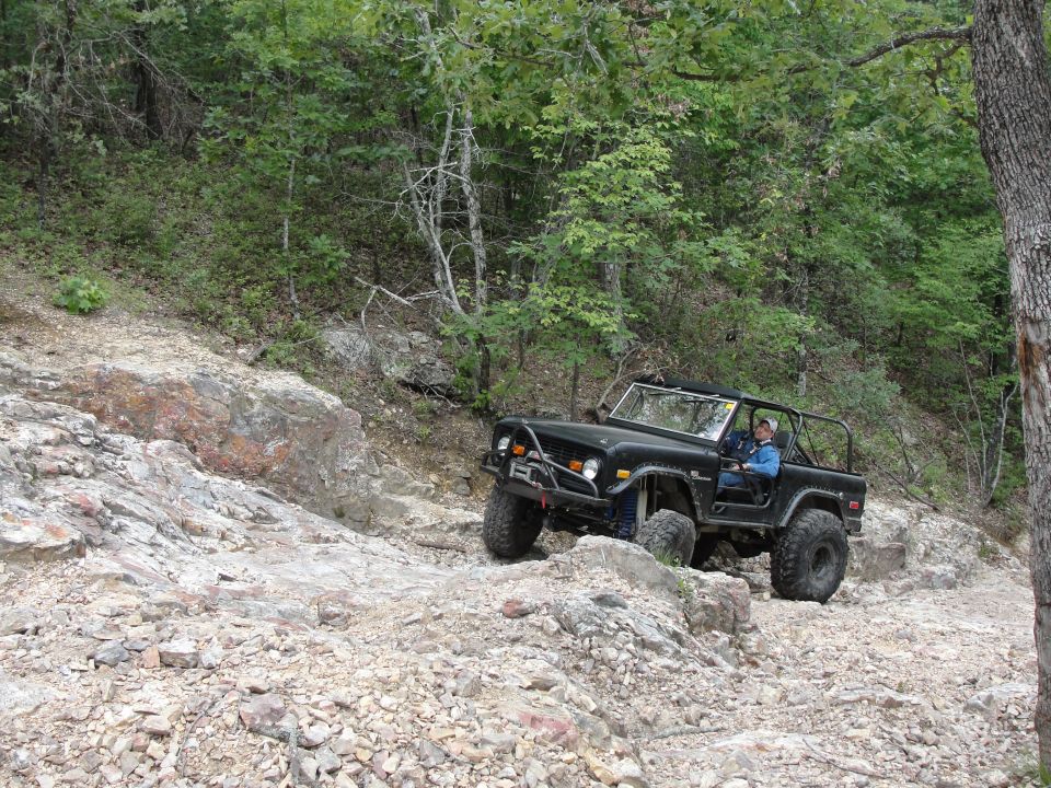 Black Ford Bronco with roll cage driving over rocks on a wooded trail