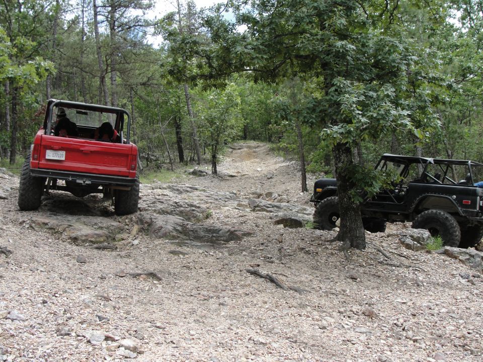 Two off-road vehicles, one red and one black, navigate a rocky, wooded trail