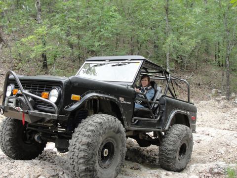 1967 Ford Mustang - A woman drives a heavily modified black Ford Bronco with large off-road tires through a wooded trail.