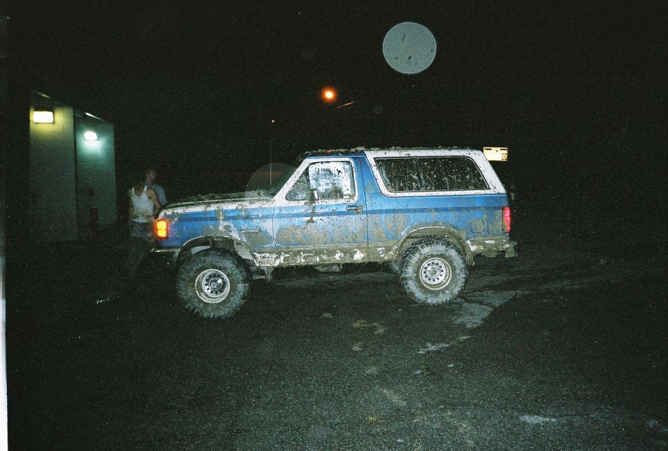 Muddy blue Ford Bronco with white top at night after off-roading, side view