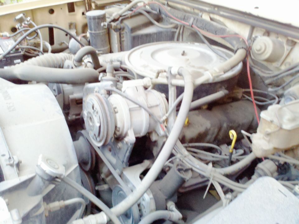 Engine bay of a beige truck, showing the V8 engine, air conditioning compressor, and fan. The engine is dusty and appears to be from the 1980s