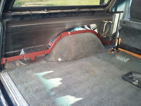 1989 Ford Bronco - The interior cargo area of an SUV shows grey carpeting and a red-painted wheel well cover.
