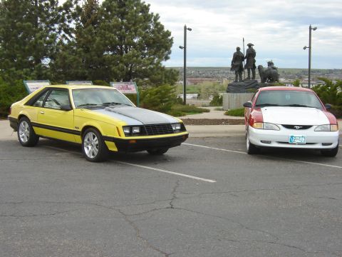 1979 Ford Mustang - A yellow 1979 Ford Mustang coupe is parked in a lot, with a red and white car to its right and a statue in the background.