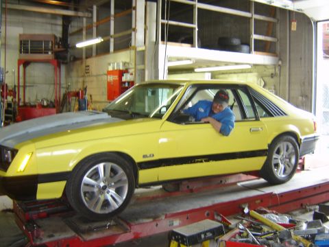 1979 Ford Mustang - A yellow 1979 Ford Mustang coupe is positioned on a red lift in a garage, with a person visible through the driver's side window.