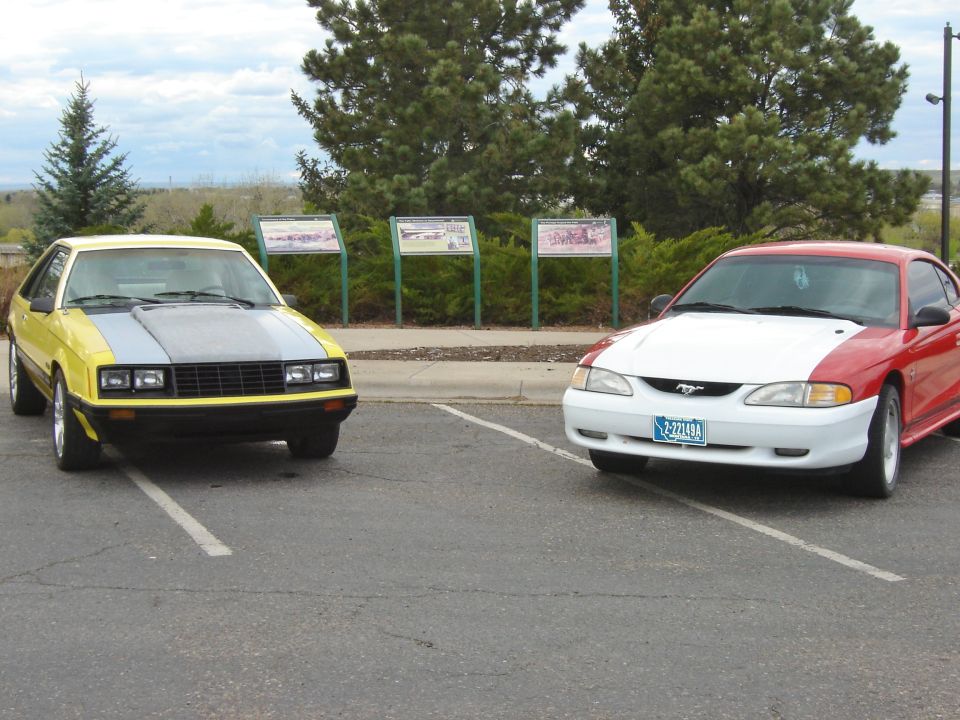 1995 Ford Mustang with red body and white hood parked next to an older yellow Mustang