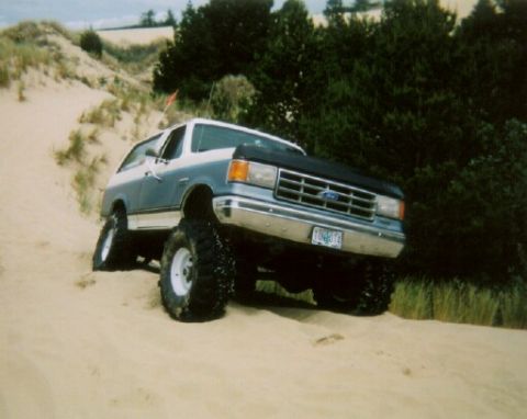 1988 Ford Bronco - A lifted Ford Bronco navigates sandy terrain with dense trees in the background.