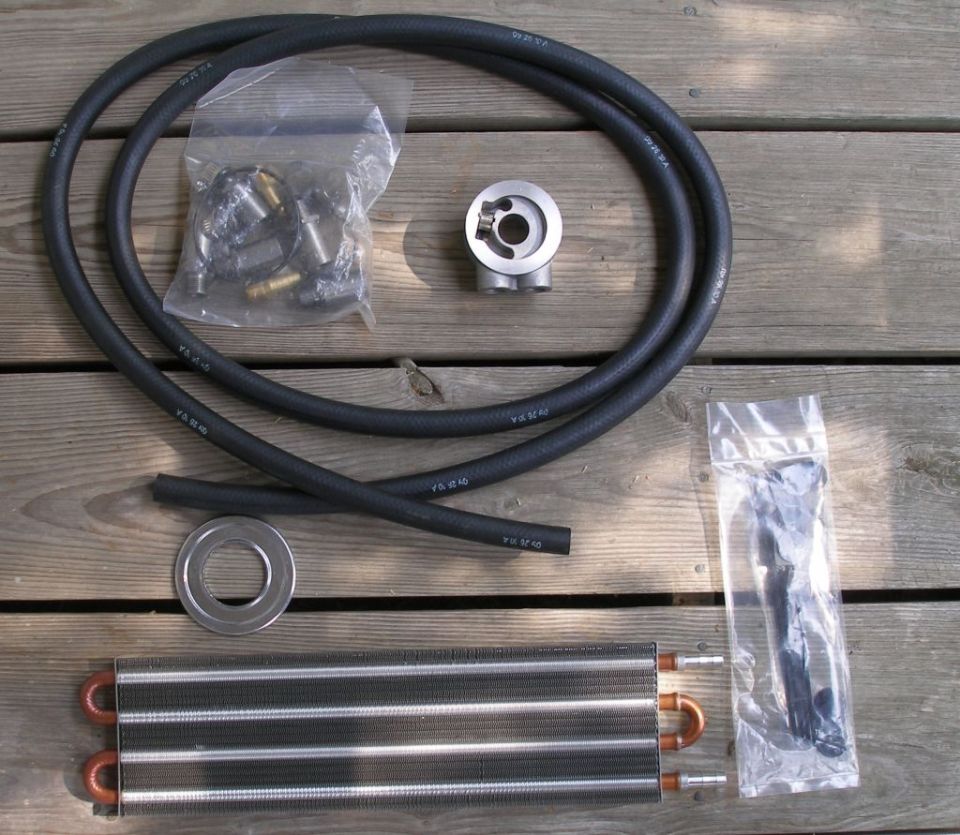 Oil cooler kit laid out on wooden planks. Includes a finned cooler, two black hoses, a metal adapter, a bag of brass fittings, and a bag of black hardware