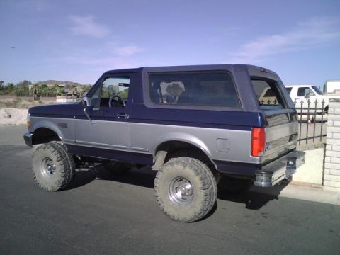 1995 Ford Bronco - A lifted Ford Bronco with a two-tone paint job is parked on a street, with another vehicle visible in the background.