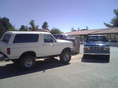 1995 Ford Bronco - A white 1995 Ford Bronco and a blue Ford F-150 are parked in front of a house on a sunny day.
