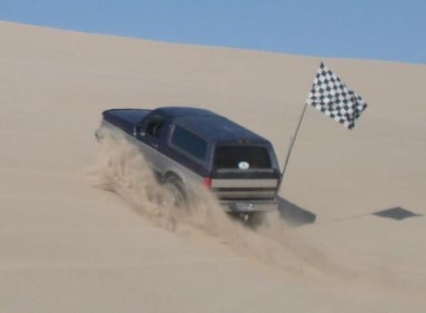 1995 Ford Bronco - A two-tone Ford Bronco navigates sandy terrain, kicking up a cloud of dust as it goes.