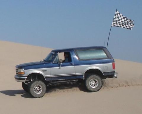 1995 Ford Bronco - A blue and silver 1995 Ford Bronco navigates a sandy terrain, a checkered flag flying high in the clear sky.