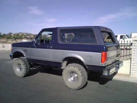 1995 Ford Bronco - This lifted Ford Bronco is parked on a street with a white SUV visible behind a fence.