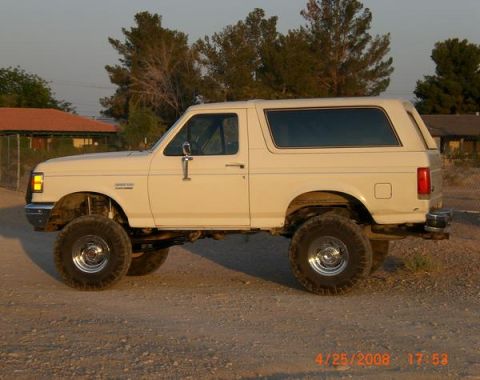 1995 Ford Bronco - A lifted white Ford Bronco is parked on a dirt road with houses in the background.