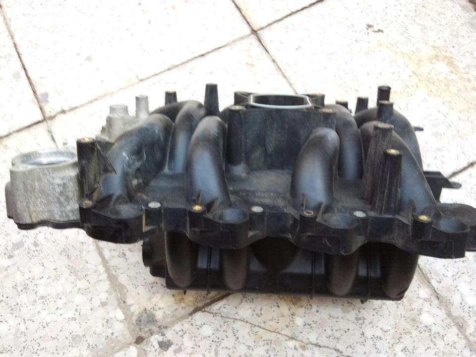 Black plastic intake manifold with silver metal component on the left, resting on a tiled surface