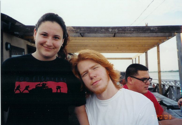 Three people stand on a deck; the woman on the left wears a black shirt with a red Foo Fighters logo