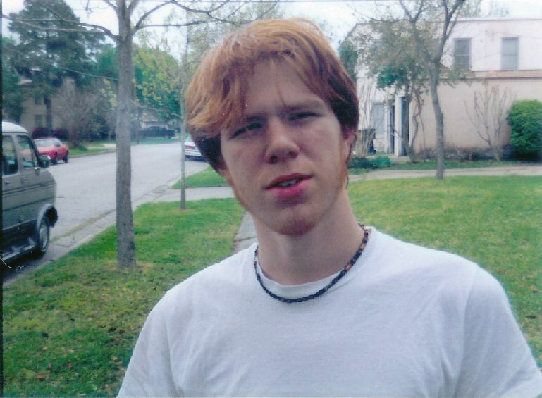 Young man with reddish hair and a white t-shirt stands on a grassy area next to a street