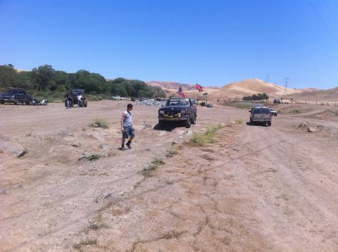 1990 Ford Bronco - A dark blue Ford F-Series truck with two flags flies down a dirt road in a dry, hilly landscape.