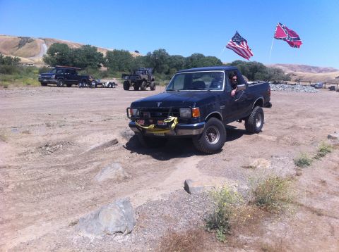 1990 Ford Bronco - A black pickup truck drives off-road with two flags flying from the bed.