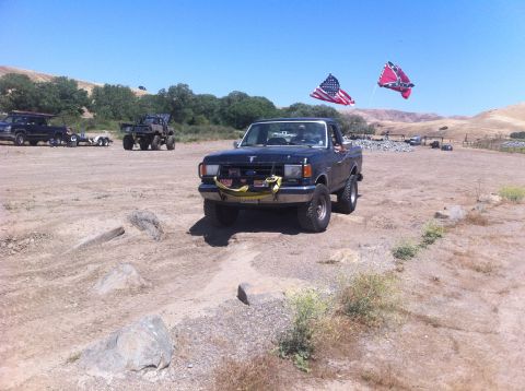 1990 Ford Bronco - A black Ford F-150 pickup truck is parked in a dusty outdoor area, with two flags flying from poles on its roof.