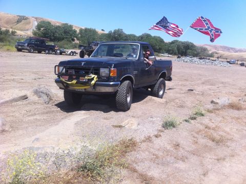 1990 Ford Bronco - A black Ford F-150 pickup truck drives off-road with two flags flying.