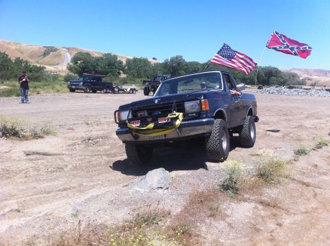 1990 Ford Bronco - Ford Bronco with flags flying on a dirt road, other vehicles visible in the background.