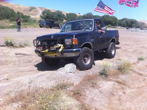 1990 Ford Bronco - A black Ford Bronco is parked on a dirt trail, with American and Confederate flags flying above.