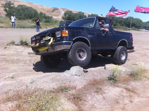 1990 Ford Bronco - Ford Bronco navigates rocky terrain under a clear sky, with flags visible in the background.