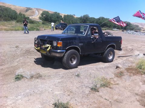 1990 Ford Bronco - A black Ford Bronco is parked on a dirt road, with two flags flying from its rear.