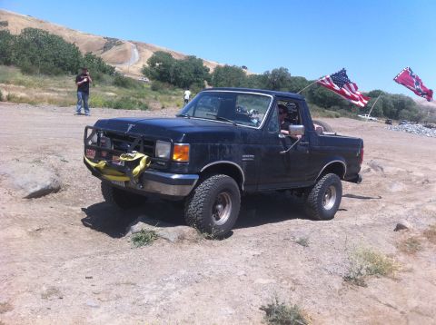 1990 Ford Bronco - A black Ford Bronco is parked on a dirt road with two flags flying in the background.