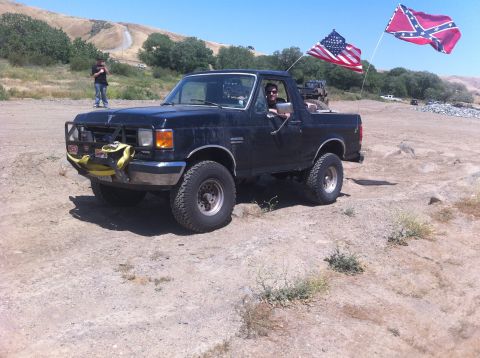 1990 Ford Bronco - Black 1990 Ford Bronco parked off-road with American and Confederate flags.