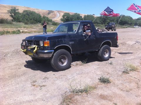 1990 Ford Bronco - A black Ford Bronco is parked off-road with two other vehicles visible in the background.