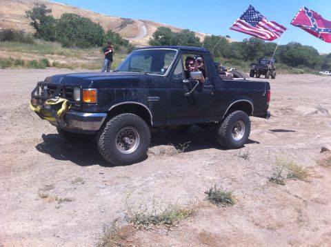 1990 Ford Bronco - A black 1990 Ford Bronco is shown off-road with American and Confederate flags flying.