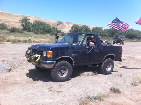 1990 Ford Bronco - Black 1990 Ford Bronco with American flag, parked off-road. Another vehicle is in the background.