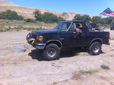 1990 Ford Bronco - A black Ford Bronco is parked off-road with a person inside and an American flag flying from the rear.