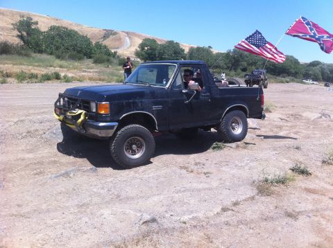 1990 Ford Bronco - A black 1990 Ford Bronco is shown off-road with American and Confederate flags flying from the rear.