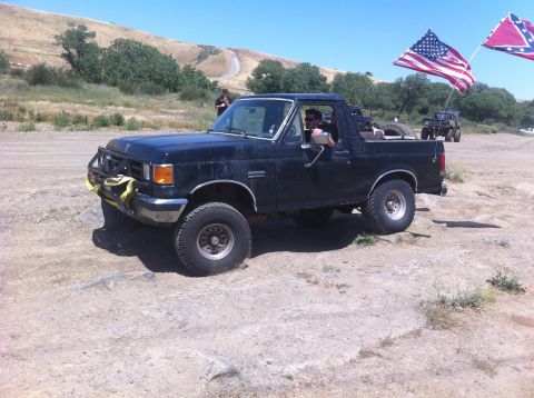 1990 Ford Bronco - Black 1990 Ford Bronco with brush guard and flags on rear, driving on dirt terrain with another vehicle in background.